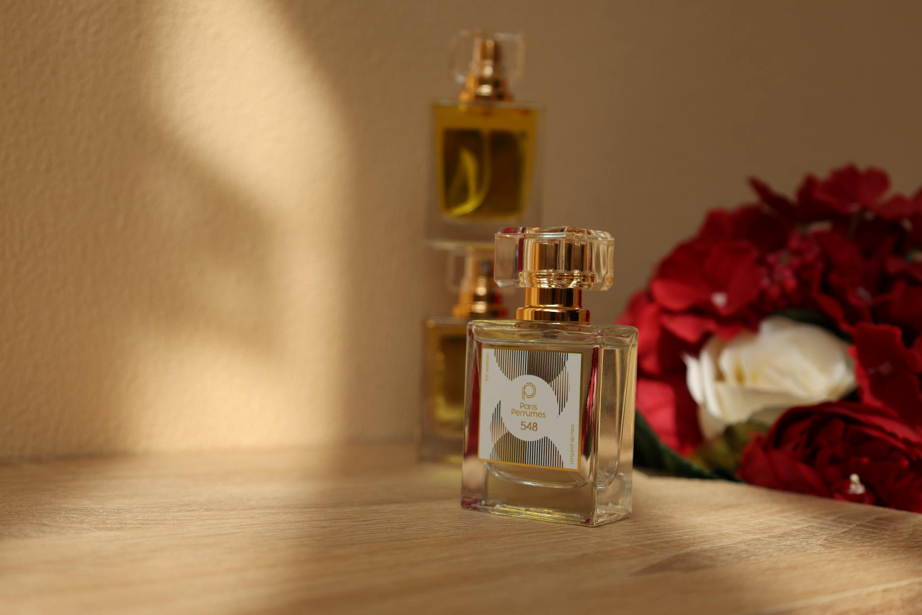 A close-up of luxurious perfume bottles beside red and white roses, elegantly lit by soft sunlight.