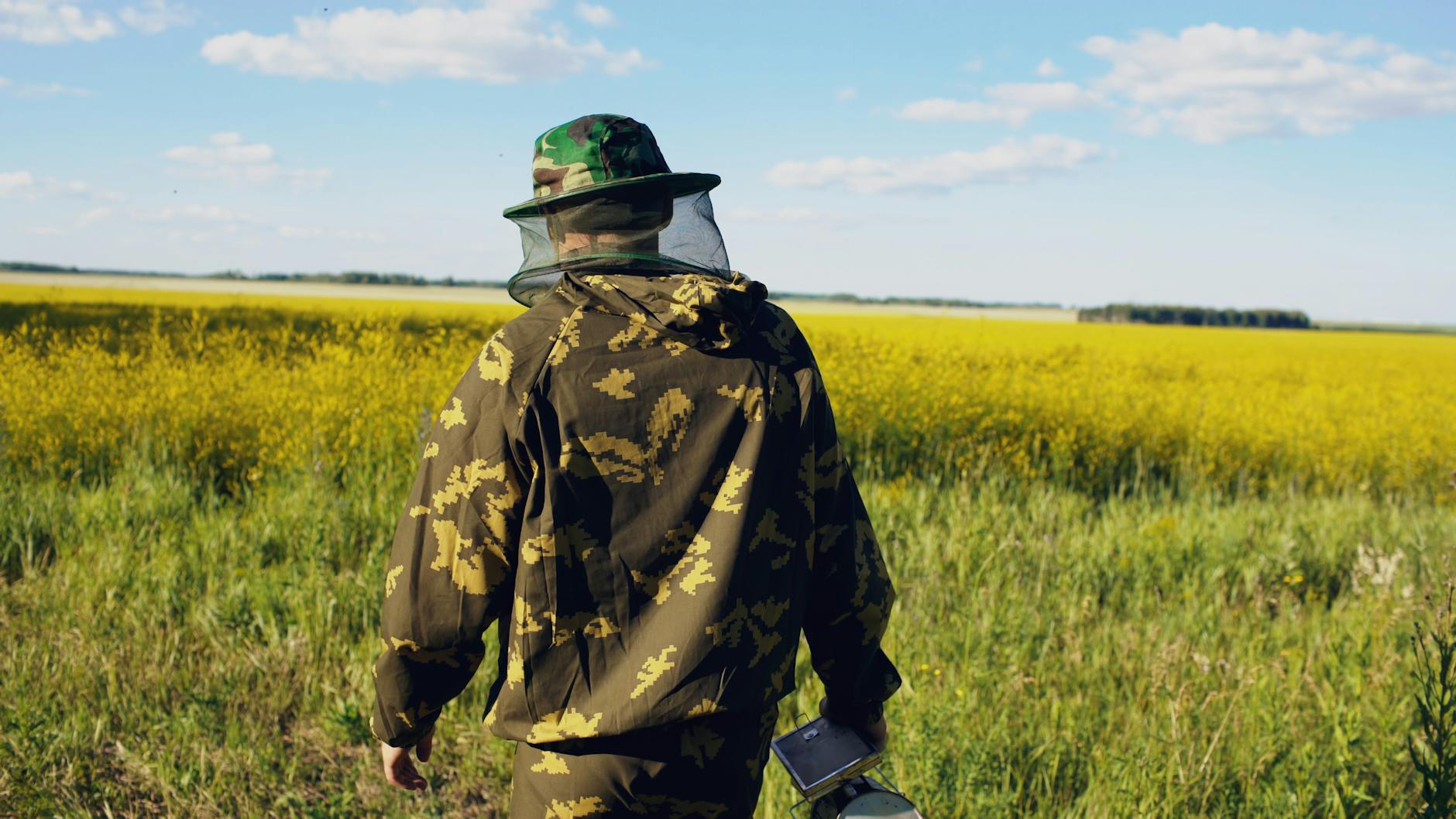 Beekeeper wearing camouflage in a lush field of yellow flowers under a bright sky.