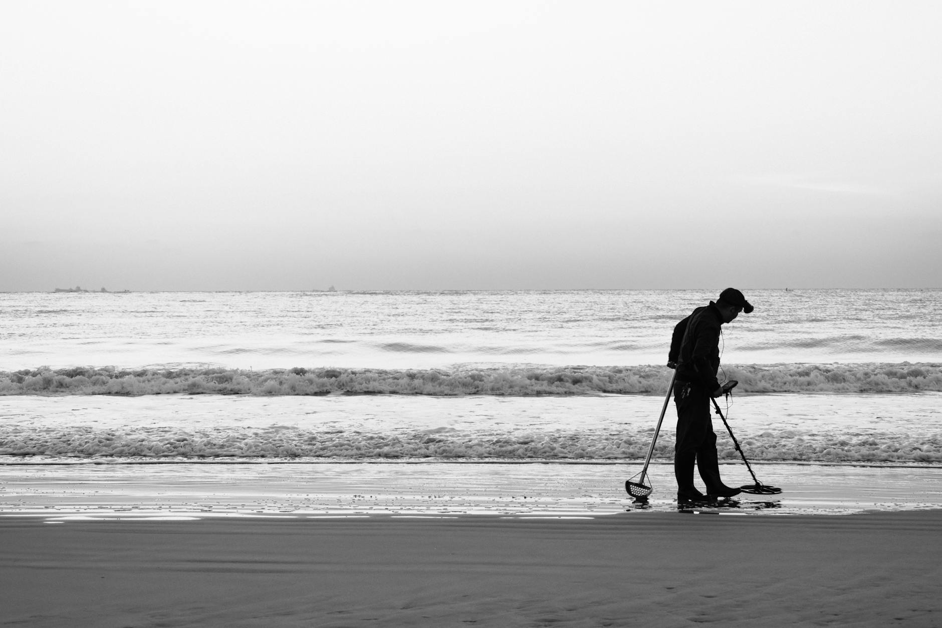 Black and white image of a silhouette of a man using a metal detector on a tranquil beach.