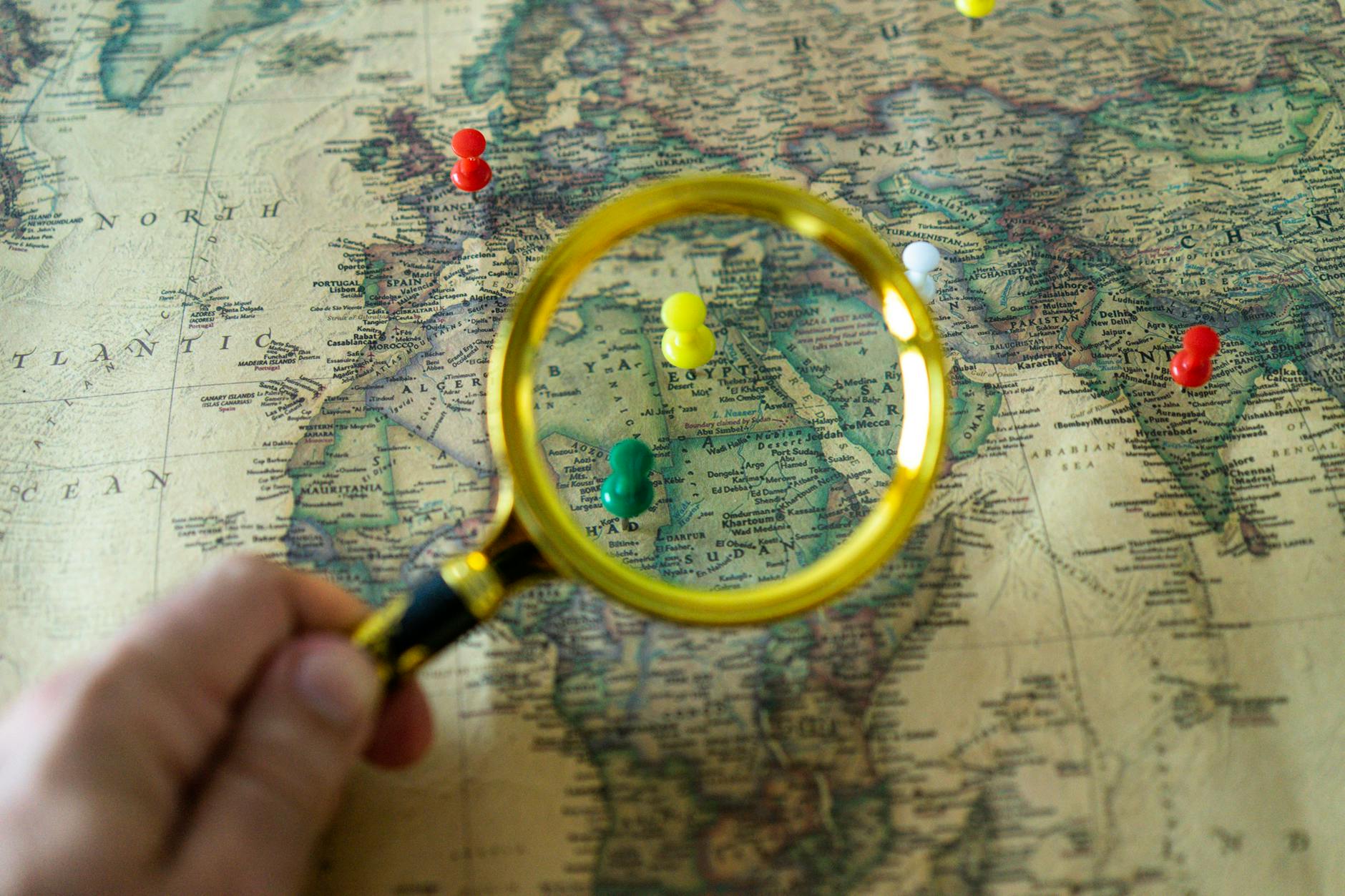 Close-up of a hand holding a magnifying glass over a world map with colorful pins indicating travel destinations.